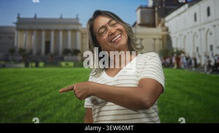 Eine Frau mittleren Alters mit Brille und gestreiftem Hemd zeigt mit dem Zeigefinger auf ein neoklassizistisches Gebäude auf einem grünen Museumsrasen, lachend und s Stockfoto