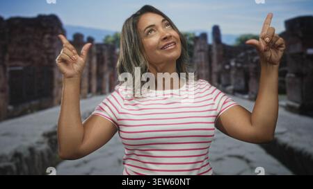 Die Frau zeigt mit den Zeigefingern nach oben auf die Steinstraße, trägt ein rosa gestreiftes Hemd und blickt nach oben; Reisewunderung. Stockfoto