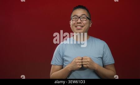 Mann, jung, mit Brille und blauem T-Shirt mit kurzen Haaren, geballten Fäusten auf der Brust in einem roten Atelier; Freude. Stockfoto