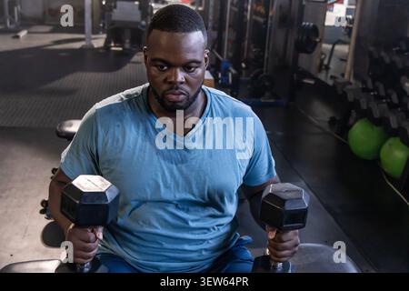 Afroamerikaner sitzt auf der Bank im Fitnessstudio und hält Hexen-Kurzhanteln, trägt schwitzendes blaues T-Shirt Stockfoto