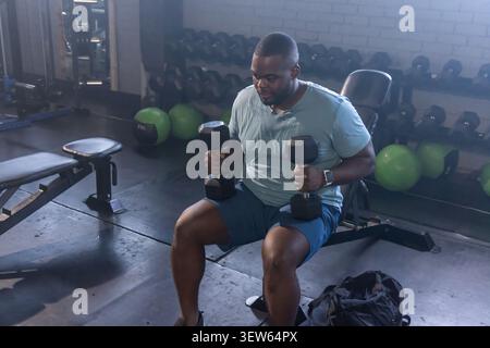 Afroamerikaner sitzt auf einer verstellbaren Hantelbank im Fitnessstudio und hält schwarze Hexen-Kurzhanteln Stockfoto