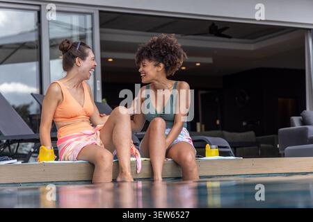Verschiedene Freundinnen in Badeanzügen sitzen zu Hause am hölzernen Poolrand und lachen mit Tassen Stockfoto