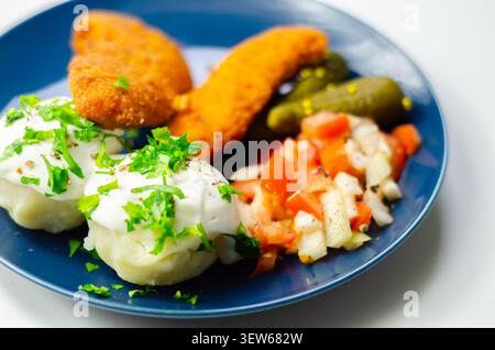 Goldene gebratene Hähnchenstangen sitzen auf einem blauen Teller mit frischer Petersilie, Gurken und cremiger weißer Sauce. Das Essen sieht appetitlich und ausgewogen aus, pe Stockfoto