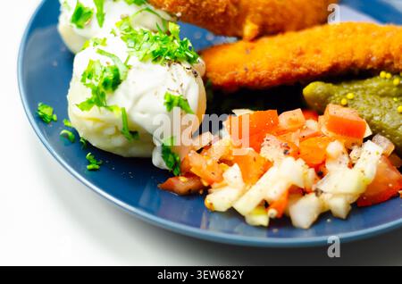 Goldene gebratene Hähnchenstangen sitzen auf einem blauen Teller mit frischer Petersilie, Gurken und cremiger weißer Sauce. Das Essen sieht appetitlich und ausgewogen aus, pe Stockfoto