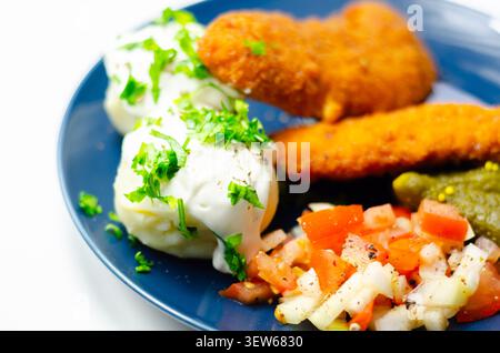 Goldene gebratene Hähnchenstangen sitzen auf einem blauen Teller mit frischer Petersilie, Gurken und cremiger weißer Sauce. Das Essen sieht appetitlich und ausgewogen aus, pe Stockfoto