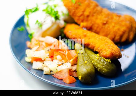 Goldene gebratene Hähnchenstangen sitzen auf einem blauen Teller mit frischer Petersilie, Gurken und cremiger weißer Sauce. Das Essen sieht appetitlich und ausgewogen aus, pe Stockfoto