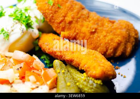 Goldene gebratene Hähnchenstangen sitzen auf einem blauen Teller mit frischer Petersilie, Gurken und cremiger weißer Sauce. Das Essen sieht appetitlich und ausgewogen aus, pe Stockfoto