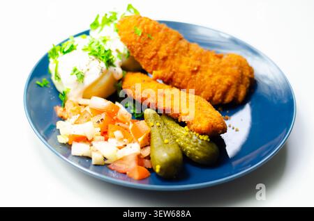 Goldene gebratene Hähnchenstangen sitzen auf einem blauen Teller mit frischer Petersilie, Gurken und cremiger weißer Sauce. Das Essen sieht appetitlich und ausgewogen aus, pe Stockfoto