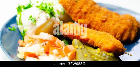 Goldene gebratene Hähnchenstangen sitzen auf einem blauen Teller mit frischer Petersilie, Gurken und cremiger weißer Sauce. Das Essen sieht appetitlich und ausgewogen aus, pe Stockfoto