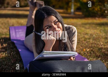 Junge Frau, die auf Gras liegt, Brainstorming macht, studiert oder schreibt in ihrem Notizblock Stockfoto