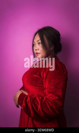 Eine junge Frau in einem roten Kleid posiert seitlich vor einem rosafarbenen Atelierhintergrund. Stockfoto