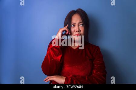 Nachdenkliche junge asiatische Frau, die verwirrt vor blauem Atelierhintergrund aussieht. Stockfoto