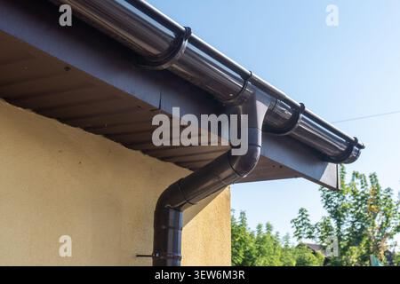 Wasser bewegt sich entlang der Dachrinne und tritt an einem hellen Tag in den fallauslauf eines Hauses ein. Bäume sind im Hintergrund sichtbar. Stockfoto