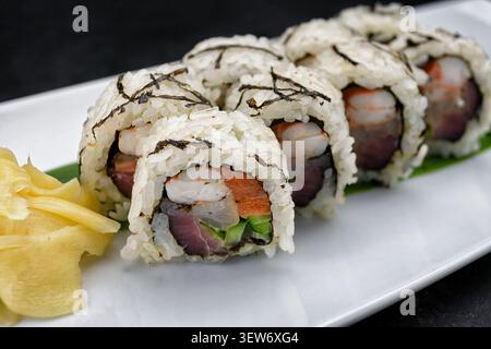 Nahaufnahme von Uamaki-Sushi-Rollen mit Garnelen, Thunfisch, Nori und eingelegtem Ingwer auf weißer Platte, dunkler Hintergrund. Restaurant mit japanischer Küche. Stockfoto