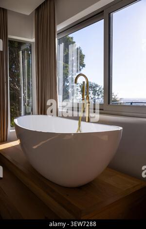 Die weiße ovale Badewanne befindet sich auf einer Holzplattform im modernen Badezimmer mit Messingarmatur an den Fenstern Stockfoto
