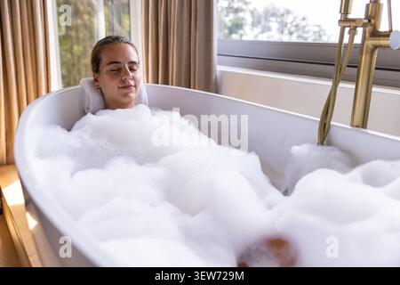 Frau, die sich in einer freistehenden Badewanne in der Nähe des Fensters liegend und sich mit dickem Schaum und Messinghahn entspannte Stockfoto