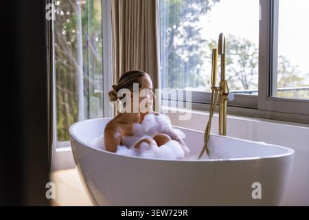 Frau, die in einer freistehenden ovalen Badewanne mit weißem Schaum sitzt, Messinghahn sichtbar, Sichtfenster Stockfoto