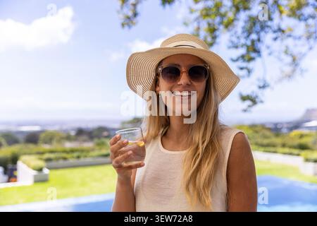 Frau mit Strohsonnenhut und Sonnenbrille, die Glas mit Zitronenscheibe im Hinterhof am Pool hält Stockfoto