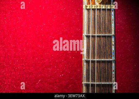Dunkles Holz Gitarrengriffbrett, metallische Saiten und Stimmstifte, leuchtend rot strukturierter Hintergrund, betont komplexe Handwerkskunst, detaillierte Nahaufnahme Stockfoto