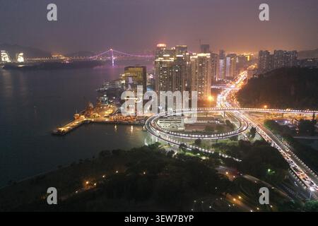 Ein nächtlicher Blick aus der Luft fängt eine hell beleuchtete Kreisrampe und die Türme am Wasser in Xiamen, China, mit Streifen von Lichtspuren im Auto, die den Fluss kurbeln Stockfoto