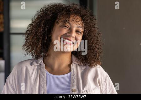 Afroamerikanerin lächelt, trägt beigefarbenes Hemd und gerippten Tank an Fensterläden im Zimmer Stockfoto