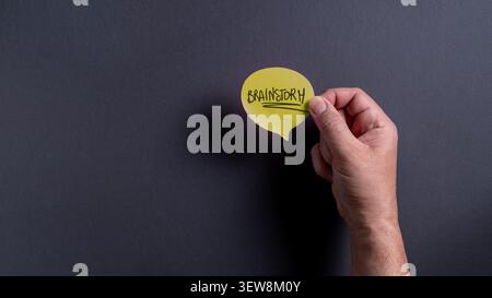 Nahaufnahme einer kaukasischen männlichen Hand, die eine gelbe Sprachblase mit handgeschriebenem Brainstorm-Text auf einem dunkelgrauen Hintergrund hält. Stockfoto