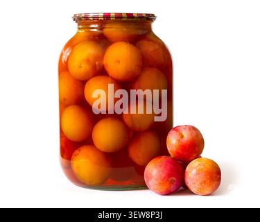 Dosenpflaumen in einem Glasgefäß auf weißem Hintergrund. Im Vordergrund stehen frische Pflaumen. Hausgemachte Obstkonserven. Stockfoto