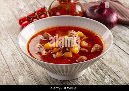 Kräftige rote Tomatenbrühe mit Fleischbällchen, Kartoffelwürfel in einer weißen Keramikschale. Rustikaler Holztisch Hintergrund mit Kirschtomaten rote Zwiebeln. Warmes Licht, Stockfoto