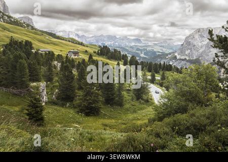 Ein Auto aus der Vogelperspektive fährt eine gewundene Bergstraße durch die Dolomiten-Landschaft mit Alpenwald, rustikalen Hütten, grünen Wiesen und Dramatten Stockfoto