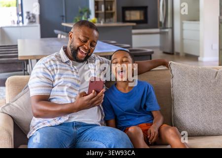 Afroamerikanischer Vater und Sohn lachen, während er zu Hause auf dem beigefarbenen Sofa auf das rote Smartphone blickt Stockfoto