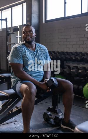 Afroamerikaner sitzt auf der Bank und hält eine Hexenhantel im Fitnessstudio. Er trägt ein hellblaues Hemd Stockfoto