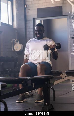 Afroamerikaner sitzt auf der Bank, trägt weißes T-Stück, hält eine Hantel und lockt sich im Fitnessstudio Stockfoto