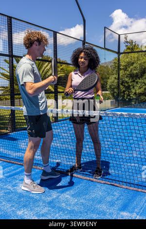 Vielfältiges Duo, das am Netz auf dem blauen Padel-Platz steht, mit schwarzen Paddeln, grünem Ball, in Sportbekleidung Stockfoto