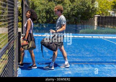 Verschiedene Freunde in sportlicher Kleidung laufen auf dem blauen Tennisplatz neben dem Netz mit Seesäcken, Telefon Stockfoto