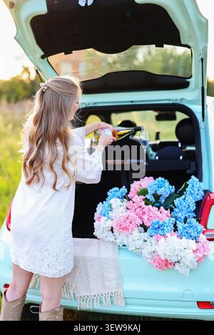 Luxuriöses Picknickkonzept im Freien mit lächelnder Frau, Sekt, bunten Blumen und Pastellfahrzeug in ruhiger ländlicher Umgebung Stockfoto
