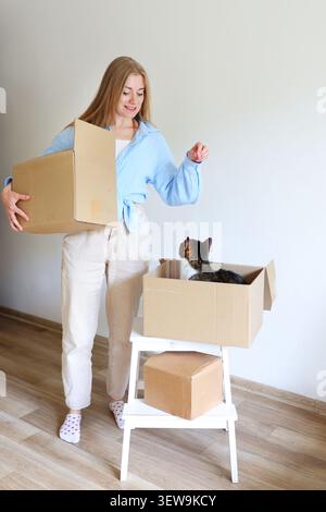 Glückliche Hausbesitzerin, die Umzugskartons trägt und mit ihrer Katze interagiert, während die Wohnung verlegt und ausgepackt wird. Stockfoto