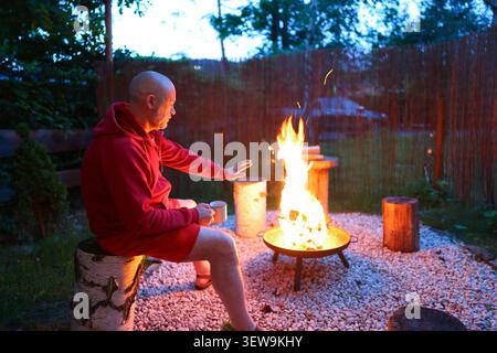 Ein Mann wärmt seine Hände am Lagerfeuer in der Abenddämmerung, genießt einen friedlichen Rückzugsort im Freien und einen gemütlichen Abend in der Natur. Stockfoto