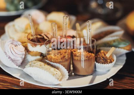 Süße Desserts glänzen auf einem rustikalen Teller. Die Glasbecher halten reichhaltige, strukturierte Leckereien mit winzigen Löffeln. Mini-Baiser und Gebäck sorgen für weiche, zarte Kontraste Stockfoto