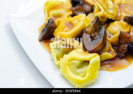 Goldene Tortellini paart mit zarten Pilzen in reichhaltiger Sauce. Ordentlich auf einer sauberen, weißen Schale aufgebracht. Soße leuchtet unter weichem, gleichmäßigem Licht. Einfach, wa Stockfoto