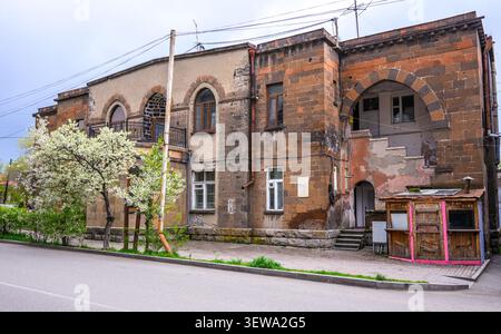 Altes Backsteingebäude mit Bogenfenstern und abblätternder Farbe. Ein Baum mit weißen Blüten steht neben der Straße Stockfoto