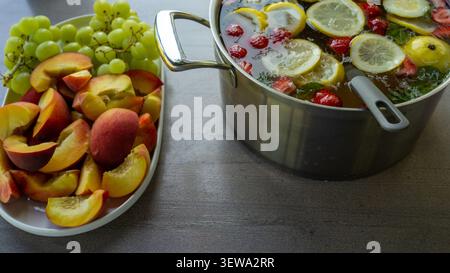 Geschlitzte Pfirsiche, grüne Trauben, Zitronen, Erdbeeren, Minze und Eis in einer Servierpfanne neben einem Obstteller auf grauem Tisch, die eine helle, gesunde Sommeressen-Stillleben mit frischen Farben und hausgemachtem Charme schaffen Stockfoto
