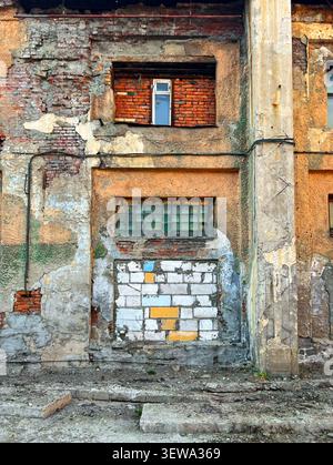 Abblätternde Putzfassade eines verlassenen Industriegebäudes mit alten blockierten Fenstern und freiliegenden Ziegeln Stockfoto