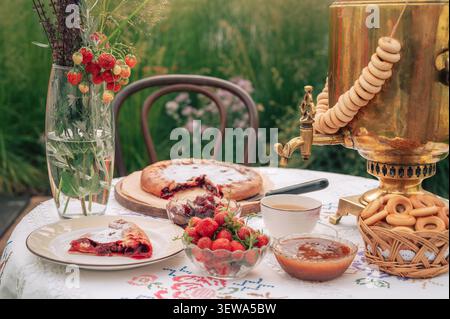 Eine rustikale Teeumgebung mit goldenem Samovar, frischen Erdbeeren, Marmelade, Kuchen und Bagels, die auf einer gestickten Tischdecke im Freien angeordnet sind. Stockfoto