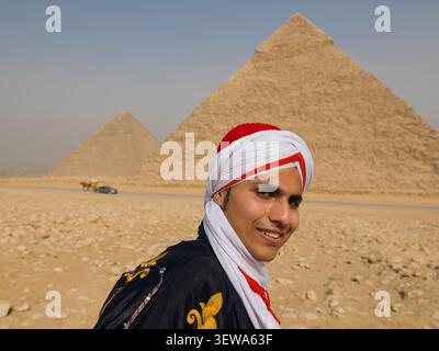 Freundlicher junger ägyptischer Mann in traditioneller Kleidung und rotem Turban, der warm vor den Pyramiden von Gizeh lächelt, ideal für Ägyptenreisen und Stockfoto