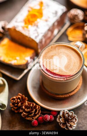Eine Glasschale Cappuccino mit herzförmiger Latte Art und rotem Puderstaub, serviert mit geschnittenem Urlaubskuchen im Hintergrund. Stockfoto