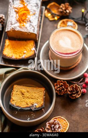 Ein Tisch mit einem warmen Cappuccino mit Herzlatte Art, einem ganzen staubigen Urlaubskuchen und einem Stück in einer dunklen Keramikschale. Stockfoto