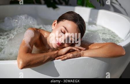 Entspannte junge kaukasische Frau, die Kopf auf den Armen ruht, während sie ein warmes Schaumbad in der Badewanne nimmt Stockfoto