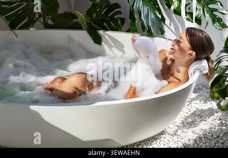 Glückliche junge kaukasische Frau, die sich in einem Sprudelbad in einem hellen, modernen Badezimmer entspannt Stockfoto