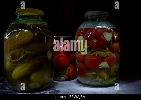 Hausgemachte eingelegte Gurken und Tomaten in Glasgläsern auf dunklem Hintergrund Stockfoto