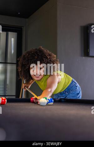 Afroamerikanerin lehnt sich auf den Billardtisch im Spielzimmer, zielt auf den Queuestift, Limettentop, Jeans Stockfoto